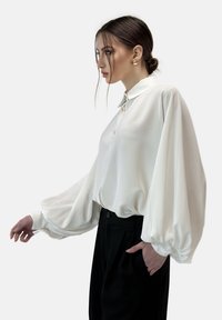 Blusa blanca y ligera con cuello button-down y mangas globo. Combinada con pantalones negros de corte ajustado. Textura suave, diseño minimalista.