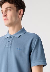 Polo de culoare bleu deschis, fabricat dintr-un material texturat, având un guler clasic și un închidere cu trei butoane, precum și un logo discret pe piept. Mâneci scurte.