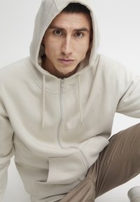 Bežová mikina na zips s veľkou kapucňou, šnúrkami a hladkou textúrou. Model pózuje sediac, oblečený v svetlohnedých nohaviciach.