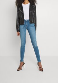 Svart lädermotorcykeljacka, vit åtsittande T-shirt, blå skinny jeans och svarta högklackade sandaler med remmar. Släta texturer och skräddarsydd passform.