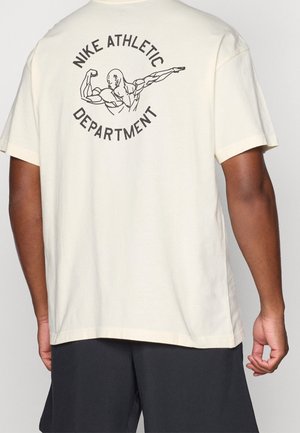 Osoba ubrana w kremową koszulkę Nike z czarną sylwetką muskularnej postaci prezentującej mięśnie oraz napisem "Nike Athletic Department" na plecach, w połączeniu z czarnymi szortami.