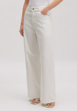 Vrouw draagt witte, hooggetailleerde jeans met wijde pijpen, hand in de zak en witte sandalen met bandjes en open teen.