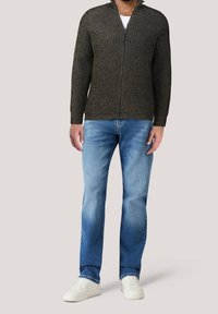 Mörkgrön, räfflad zip-up-tröja med hög krage, bärs över en vit skjorta, tillsammans med ljusblå jeans och vita sneakers.