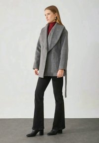 Cappotto grigio in misto lana con colletto ampio, design avvolgente e fiocco laterale, abbinato a pantaloni neri svasati e stivali, su uno sfondo neutro.