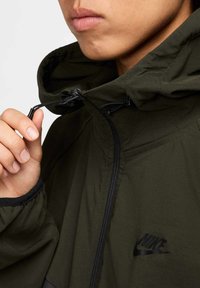 Grön huva-jacka med en slät yta, svart dragkedja och logotyp. Modellen justerar snörningen vid halsen.