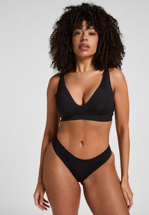 Femme aux cheveux bouclés portant un soutien-gorge noir et une culotte taille haute assortie, debout devant un fond gris clair uni.