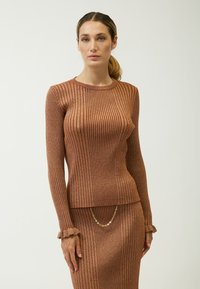 Rippendes, langärmeliges Oberteil in einem warmen, metallischen Braunton. Weiche Textur mit gefransten Manschetten. Kombiniert mit einem passenden Rock, der mit einem goldenen Kettendetail versehen ist.