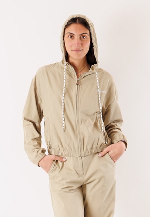 Donna che indossa una giacca beige con cappuccio e zip e pantaloni abbinati, con le mani nelle tasche, in piedi davanti a uno sfondo chiaro uniforme.