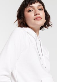 Sudadera blanca con cordón, que presenta un bolsillo frontal y detalle de texto. Textura suave, corte relajado y puños acanalados.