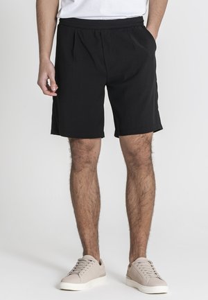 Mann trägt schwarze knielange Shorts mit Seitentaschen und beige Freizeitsneakers, steht vor einem einfarbigen hellen Hintergrund.