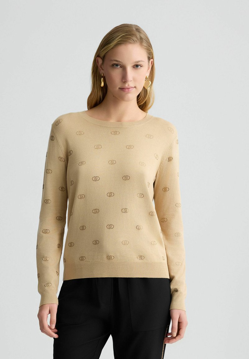 Maglione lavorato a maglia beige con un motivo logo tonale a tutta superficie, collo rotondo e maniche lunghe, abbinato a pantaloni neri.
