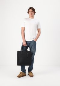 Bolso tote de cuero negro con dos asas en la parte superior, forma rectangular, textura suave y diseño minimalista. Lleva puesto alguien con atuendo casual.