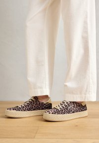 Personne portant un pantalon en velours côtelé blanc et des baskets à plateforme à imprimé léopard, debout sur un sol en bois.