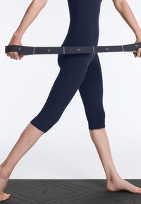 Personne en leggings bleu marine étirant une bande de résistance Pilates numérotée sur un tapis d'exercice noir, pieds nus sur le tapis.