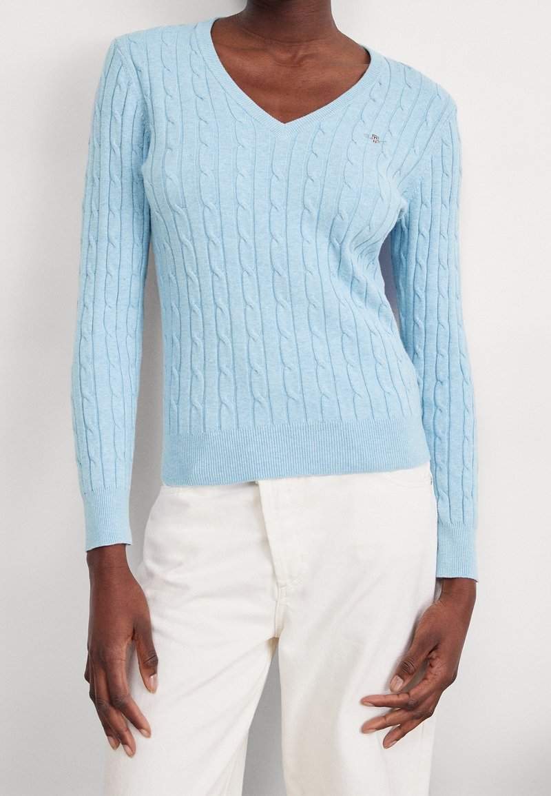 Kobieta ubrana w jasnoniebieski sweter w warkocze z dekoltem w serek i kremowe spodnie, stojąca na tle jasnego, jednolitego tła.