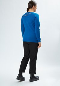 Maglione lavorato a maglia blu con dettagli in pizzo sulla schiena, abbinato a pantaloni neri e stivaletti neri chunky, mostrato di spalle.