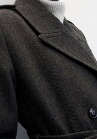 Manteau en laine à motif chevrons sombre avec un col cranté, doté d'une fermeture à boutons et d'une ceinture à nouer, porté sur un col roulé clair.