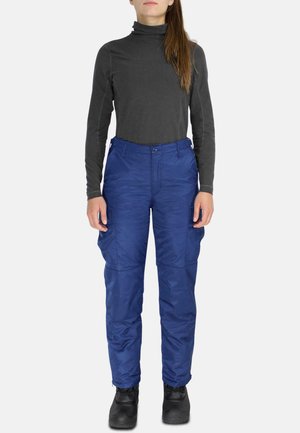 Person, die einen schwarzen Langarm-Rollkragenpullover, blaue Cargohosen mit Taschen und schwarze Stiefel trägt, steht vor einem schlichten hellen Hintergrund.