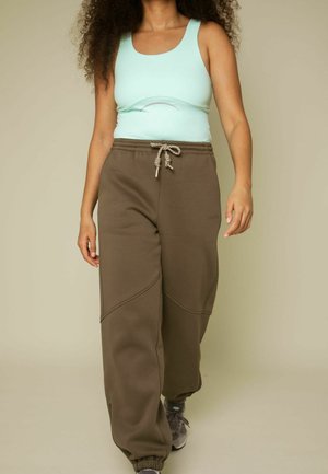 Frau trägt ein hellblaues ärmelloses Top und lockere braune Jogginghose mit Kordelzug und geht vor neutralem Hintergrund.