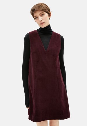 Burgunderfarbendes Corduroykleid mit tiefem V-Ausschnitt und seitlichen Taschen, getragen über einem schwarzen Rollkragenpullover. Strukturierter Stoff mit vertikalen Rillen.