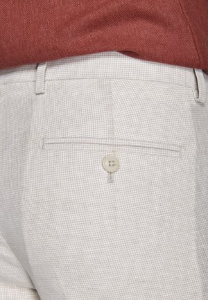 Nahaufnahme beiger karierter Hosen mit einer mit Knöpfen versehenen Gesäßtasche, getragen zu einem in die Hose gesteckten rotbraunen, strukturierten Hemd.