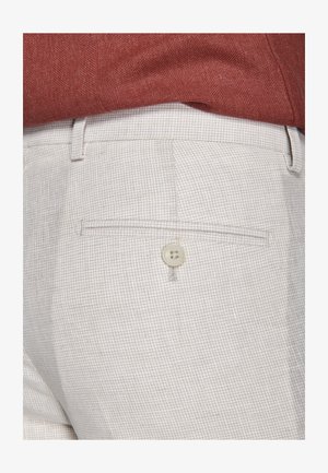 Nahaufnahme beiger karierter Hosen mit einer mit Knöpfen versehenen Gesäßtasche, getragen zu einem in die Hose gesteckten rotbraunen, strukturierten Hemd.