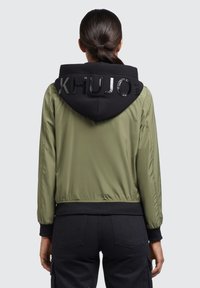 Jachetă bomber verde măslinie cu glugă neagră. Prezintă litere lucioase "KHUJO" pe glugă și manșete și tiv ribbate. Textură netedă.