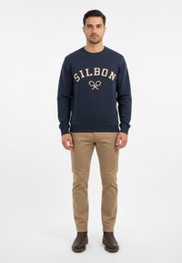 Sudadera marinera con "SILBON" y un emblema en beige. Combinada con chinos color beis y botas de tobillo marrones. Fondo sencillo.