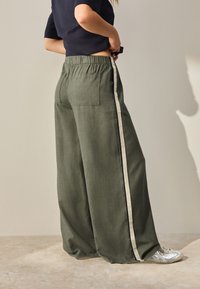 Wide-legged, green fabric pants with a textured finish, featuring a cream side stripe and an elastic waistband. One back pocket visible.
