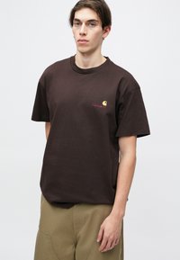 Brown cotton t-shirt with a relaxed fit, short sleeves, crew neckline, and a small Carhartt logo in yellow and red on the left chest.