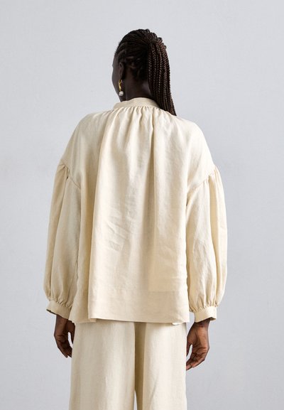 Blouse en lin crème à manches bouffantes avec un col froncé, associée à un pantalon en lin doux assorti ; texture lisse, coupe décontractée.