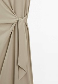 Tissu beige noué à la taille, créant des plis drapés et une ceinture lâche pendante sur le côté droit.