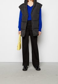 Doudoune sans manches noire à col montant, associée à un pull bleu à col en V, un pantalon noir à jambes larges et un sac à main jaune. Des chaussures noires complètent la tenue.