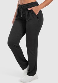 Vrouw in zwarte joggers met elastische tailleband en trekkoord, met één hand in de zak, en witte sneakers tegen een effen achtergrond.