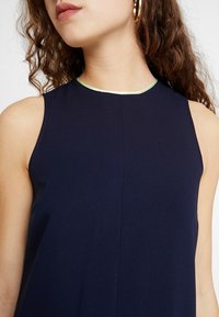 Femme portant un haut sans manches bleu foncé avec une bordure vert clair au niveau du col et de grandes boucles d'oreilles en anneaux, cheveux bouclés visibles sur un côté.