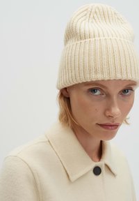 Cappello berretto in maglia color crema con texture a coste, indossato da un modello con un cappotto chiaro con un bottone. Materiale morbido e caldo con una vestibilità aderente.