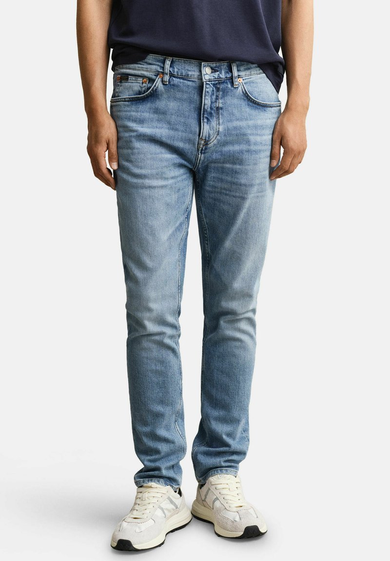 Hombre con vaqueros ajustados azul claro, camisa oscura metida por dentro y zapatillas blancas, de pie frente a un fondo liso.