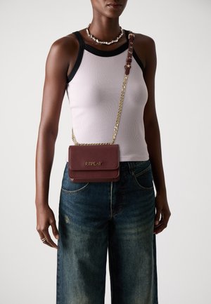 Bordeaux leren crossbody tas met een gouden schouderriem, voorzien van een klep sluiting en een geperst "REPLAY" logo. Draagt met een strakke top en denim jeans.