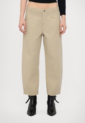 Pantalon beige droit porté avec des bottines noires, court à la taille dévoilant le ventre, et un haut noir à manches longues.