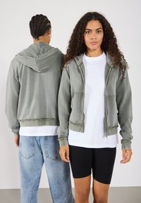Hoodie verde a coste con zip frontale, cappuccio con coulisse e polsini a costine. Indossato sopra una maglietta bianca e abbinato a jeans blu chiaro e pantaloni neri.