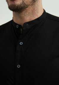 Chemise noire à col mandarin. Présente des boutons bleus et un tissu texturé. Détail de la doublure à motif cachée visible au niveau du col.