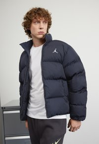 Mornarsko modra puhovka z visokim ovratnikom, oblazinjeno oblikovanje in belo Jordanovo logo na levem prsnem delu; kombinirana z belo majico in sivohlačami.