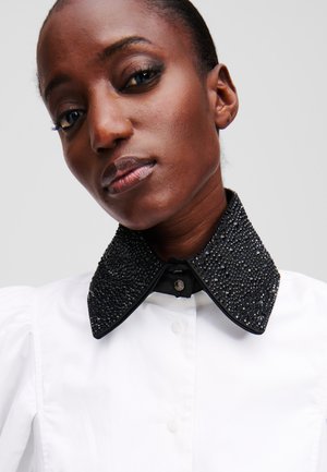 Col en perles noires sur une chemise blanche, présentant un design texturé avec des clous et une fermeture à bouton. Tissu lisse avec des manches bouffantes structurées.