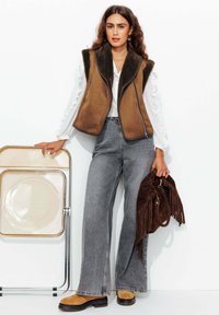 Gilet en fausse fourrure brun, blouse blanche à manches longues avec volants, jean gris à jambe large, sac à franges en suède brun, bottes à talons épais brunes, chaise minimaliste.