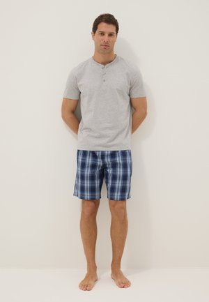 Mann steht barfuß an einer Wand und trägt ein hellgraues Henley-Shirt mit kurzen Ärmeln und blaue karierte Shorts.