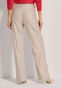 Beige, wijde broek met een zachte textuur, voorzien van twee achterzakken. Gepaard met lichtgekleurde schoenen. Achteraanzicht getoond.