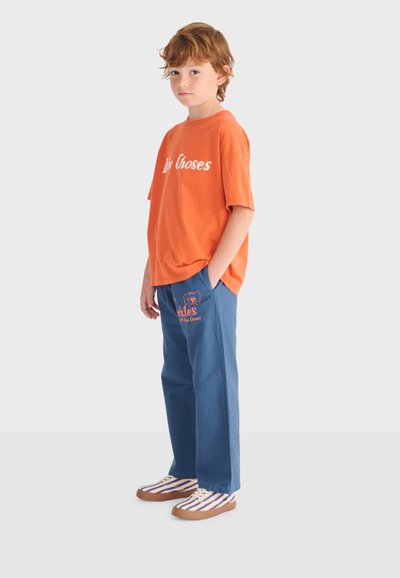 Boy with red hair stands with one hand in pocket, wearing orange T-shirt with text, blue pants with graphic, and striped sneakers on white background.