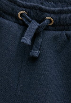 Laivastonsiniset verkkarit, joissa on leveä vyötärönauha ja paksut vetonarut, jotka kulkevat metallisten silmukoiden läpi, joihin on merkitty "Joules".