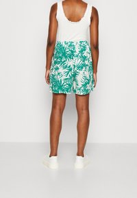 Zielone szorty w wzór palmowych liści, z elastycznym pasem i kieszeniami, zestawione z białą koszulką na ramiączkach oraz białymi trampkami.