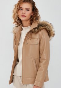 Tan faux leather jacket with a fur-lined hood, featuring zippered pockets, smooth texture, and a fitted design.